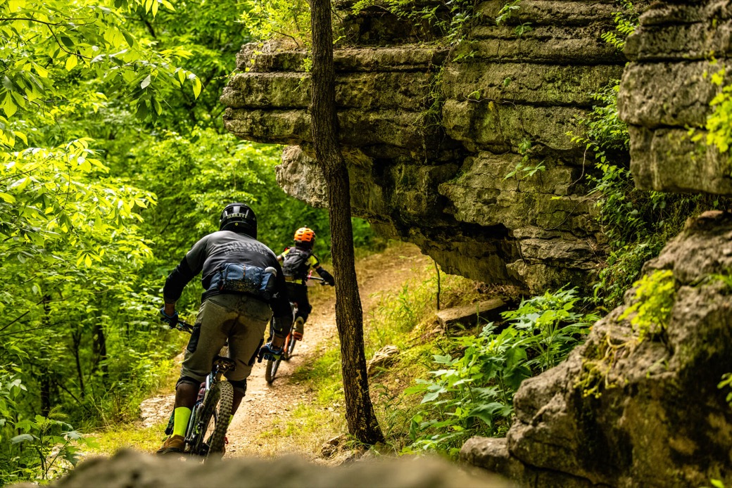 Two mountain bikers on a wooded trail beneath a limestone bluff